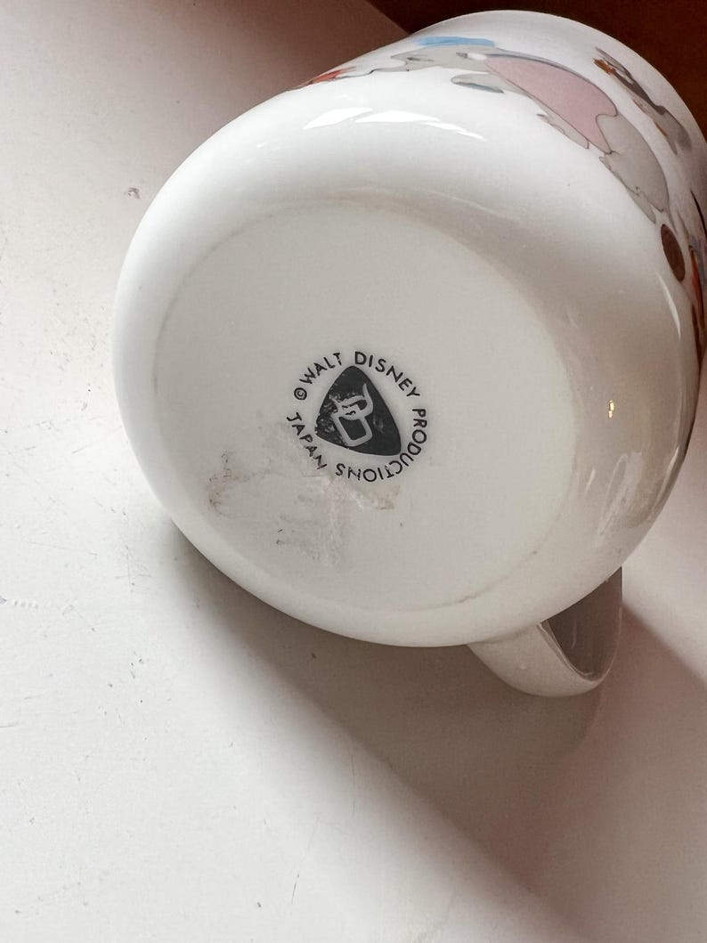 May include: White ceramic mug with a cartoon illustration. The base of the mug features the text "WALT DISNEY PRODUCTIONS JAPAN SHOW". The mug has a handle and a rounded shape. The mug is likely vintage.