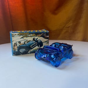 Puede incluir: Un coche de juguete de cristal azul y su caja. El coche tiene un diseño detallado con ruedas y parabrisas visibles. La caja presenta una ilustración vintage de un coche similar con gente dentro, ambientada en un entorno costero.