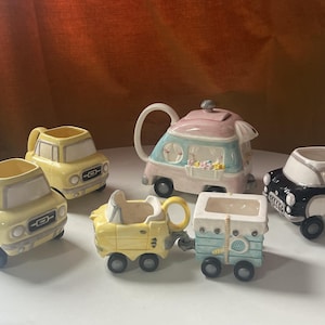 May include: A set of six ceramic teacups and teapot shaped like vintage cars. The teapot is a pink and blue camper van, and the teacups are yellow, blue, and black cars.