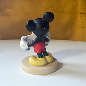 May include: A Mickey Mouse figurine, viewed from the back, stands on a beige circular base. Mickey is wearing red shorts, black legs, yellow shoes, white gloves, and a black torso and head. He is holding a light blue object.