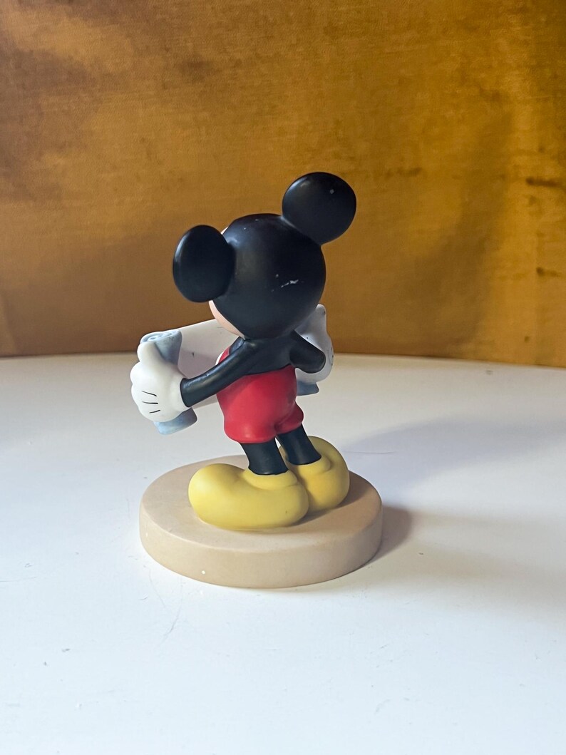 May include: A Mickey Mouse figurine, viewed from the back, stands on a beige circular base. Mickey is wearing red shorts, black legs, yellow shoes, white gloves, and a black torso and head. He is holding a light blue object.