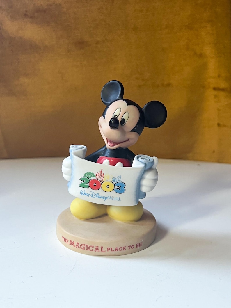May include: A Mickey Mouse figurine holding a scroll that reads "2003 Walt Disney World." The figurine has black ears, a red shirt, and yellow shoes. The base reads "THE MAGICAL PLACE TO BE!"