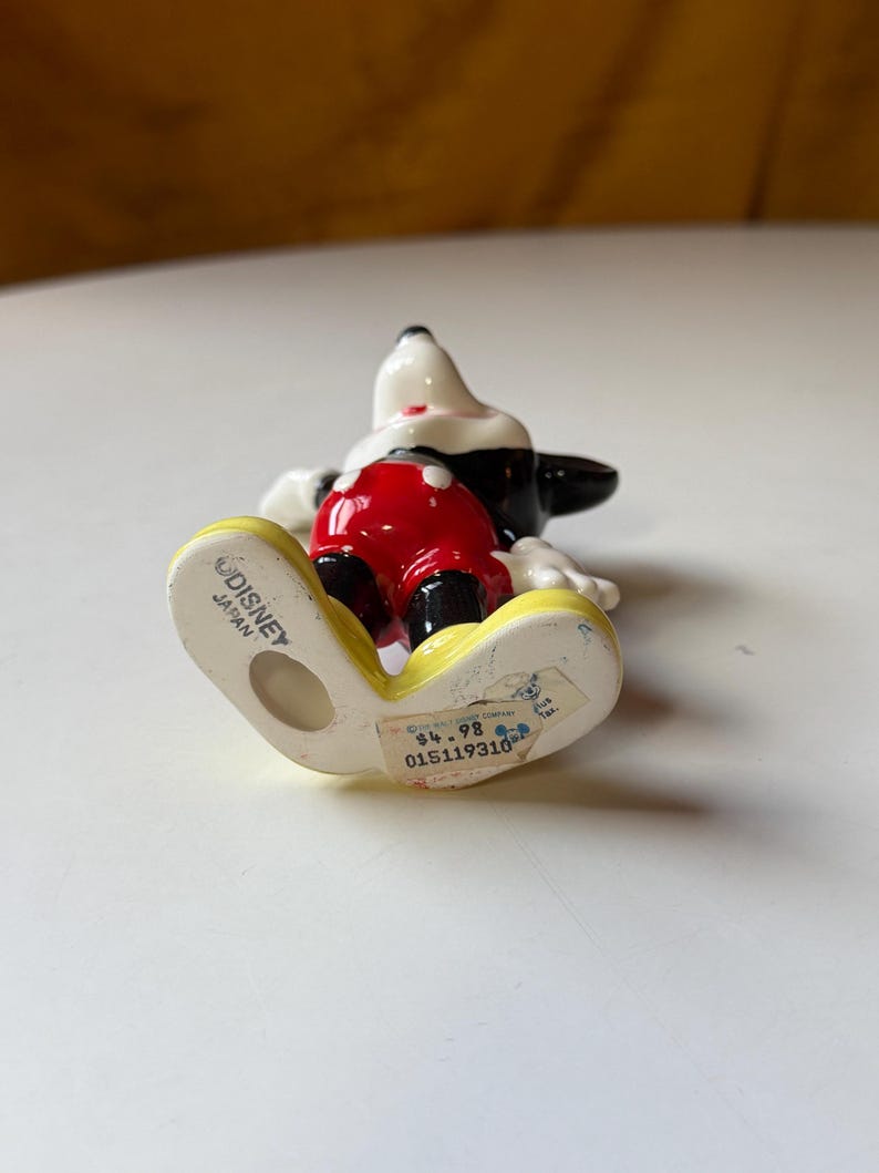 Puede incluir: Una figura de cer&aacute;mica de Mickey Mouse, con cabeza y orejas negras, guantes blancos, pantalones cortos rojos y zapatos amarillos. La base est&aacute; marcada "Disney Japan".