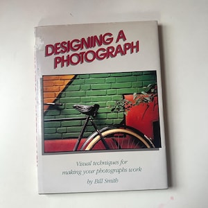 Puede incluir: Libro titulado "Designing a Photograph" con una imagen de bicicleta en la portada. La portada presenta una bicicleta negra con una rueda roja, sobre un muro de ladrillos verdes y una estructura roja. También está presente el texto "Visual techniques for making your photographs work by Bill Smith".