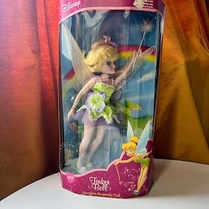 Könnte beinhalten: Eine Tinker Bell Porzellan-Andenkenpuppe in einer durchsichtigen Plastikbox. Die Puppe hat blondes Haar, grüne und lila Kleidung und einen Zauberstab. Die Box hat einen rosa Deckel mit dem Disney-Logo und den Worten "Tinker Bell" und "Porcelain Keepsake Doll".