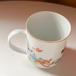 May include: White ceramic mug with a gold rim and a curved handle. The mug features colourful cartoon illustrations of Donald Duck, Pluto, and a red bird. The mug is likely for beverages.