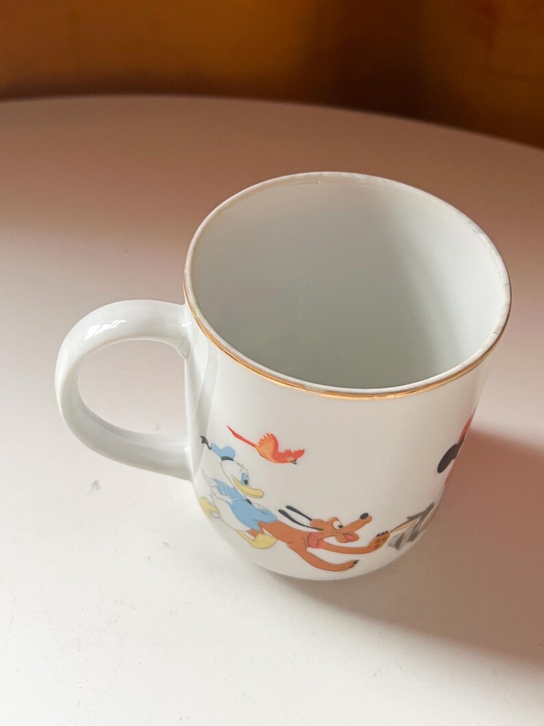 May include: White ceramic mug with a gold rim and a curved handle. The mug features colourful cartoon illustrations of Donald Duck, Pluto, and a red bird. The mug is likely for beverages.