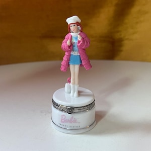 May include: A white porcelain trinket box with a pink and blue Barbie doll figurine on top. The box has a silver band around the middle and says "Barbie" in pink letters and "Groovy Sixties" in black letters.