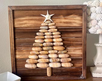 May include: A wooden framed Christmas tree made of seashells, with a white starfish on top. The tree is mounted on a wooden plank background. A white square bowl and a lamp with a seashell shade are also visible.
