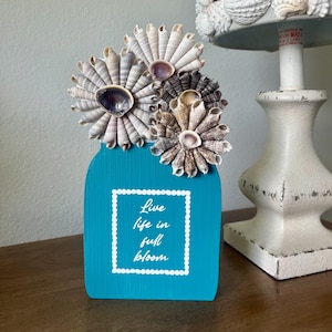 May include: A turquoise wooden vase with three seashell flowers. The vase has a white framed text that reads "Live life in full bloom." A seashell-covered lamp is in the background. The vase is on a wooden surface.