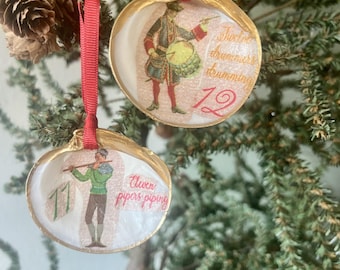 May include: Two Christmas ornaments made from seashells, each with a gold-painted rim and a red ribbon for hanging. One features a drummer and the number 12, the other a piper and the number 11. The text reads "Twelve drummers drumming" and "Eleven pipers piping".
