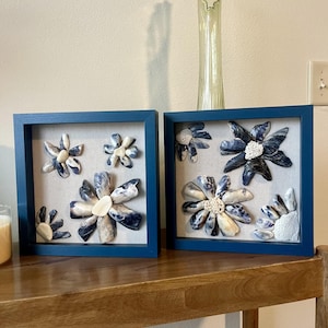 May include: Two framed floral art pieces. Each features a blue frame, a neutral linen background, and seashell flowers in shades of blue, white, and cream. The artwork is displayed on a wooden surface.