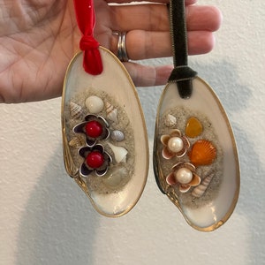 May include: Two seashell ornaments with red, white, and orange beads and seashells inside. The ornaments are attached to red and green velvet ribbon.