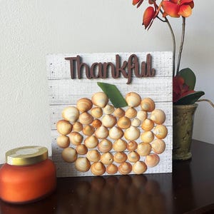 May include: A white wooden sign with the word "Thankful" in brown letters. The sign is decorated with a pumpkin made of seashells.