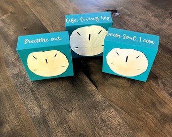 May include: Three teal blue wooden blocks with white text and a sand dollar design. The blocks read "life, living by", "breathe out", and "ocean soul: I can".