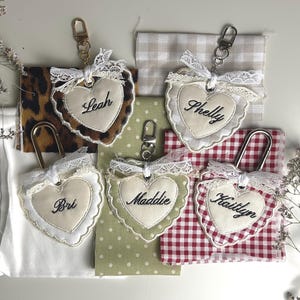 May include: Five heart-shaped fabric keychains with metal lobster clasps. Each keychain features a cream-coloured centre embroidered with the names Leah, Shelly, Bri, Maddie, or Kaitlyn, surrounded by lace trim and various patterned fabric backings.