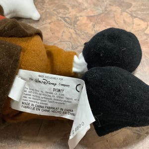 Puede incluir: Un peluche con cabeza negra, guantes blancos y abrigo marr&oacute;n. El juguete tiene una etiqueta que dice "MADE EXCLUSIVELY FOR The Walt Disney Company". La etiqueta tambi&eacute;n incluye el logotipo de Disney e informaci&oacute;n de fabricaci&oacute;n.