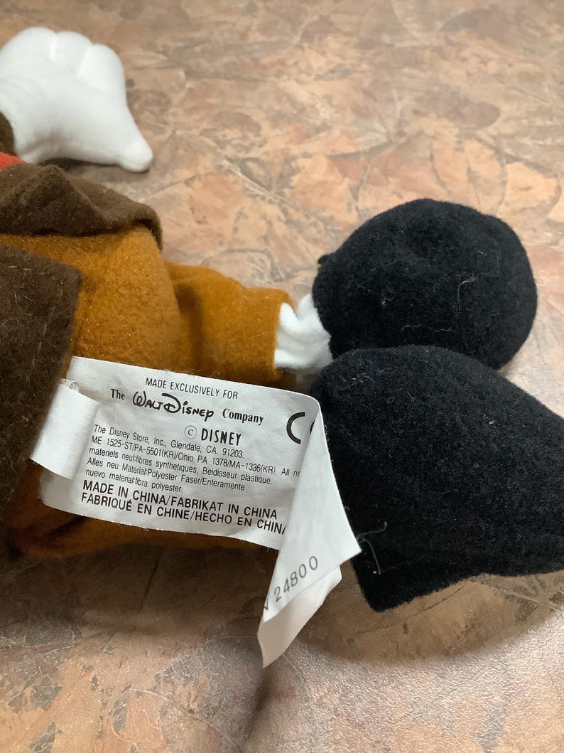 Puede incluir: Un peluche con cabeza negra, guantes blancos y abrigo marr&oacute;n. El juguete tiene una etiqueta que dice "MADE EXCLUSIVELY FOR The Walt Disney Company". La etiqueta tambi&eacute;n incluye el logotipo de Disney e informaci&oacute;n de fabricaci&oacute;n.