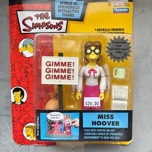 May include: A plastic action figure of Miss Hoover from The Simpsons, wearing a pink skirt, yellow blouse, and glasses. The figure is packaged in a colorful box with the words "Gimme! Gimme! Gimme!" on a sign and "Miss Hoover" on the front. The box also includes a small playset with a sign that reads "45 seconds 'til pencils down."