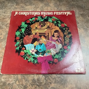 May include: Vintage vinyl record album cover for "A Christmas Music Festival." The cover features a family gathered around a fireplace, surrounded by a Christmas wreath. Presents are under the wreath, and the album cover is red.