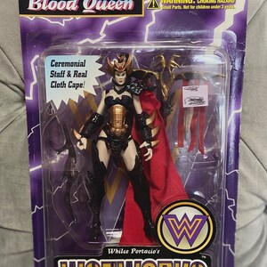 May include: A plastic package containing a Blood Queen action figure from the Wetworks Ultra-Action Figures line. The figure is a female warrior with a black and gold armor, a red cape, and a staff. The package includes the text "Ceremonial Staff & Real Cloth Cape!" and "Whilce Portacio's Wetworks Ultra-Action Figures."