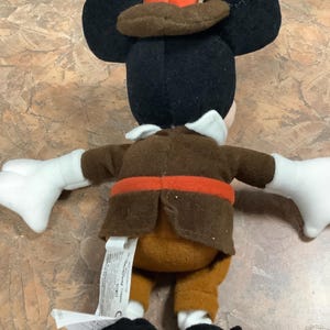 Puede incluir: Un peluche de Mickey Mouse con un sombrero y una chaqueta marr&oacute;n de peregrino con un cintur&oacute;n naranja. El juguete tiene orejas negras, una cabeza negra, guantes blancos y zapatos negros. El juguete est&aacute; sobre una superficie marr&oacute;n claro.