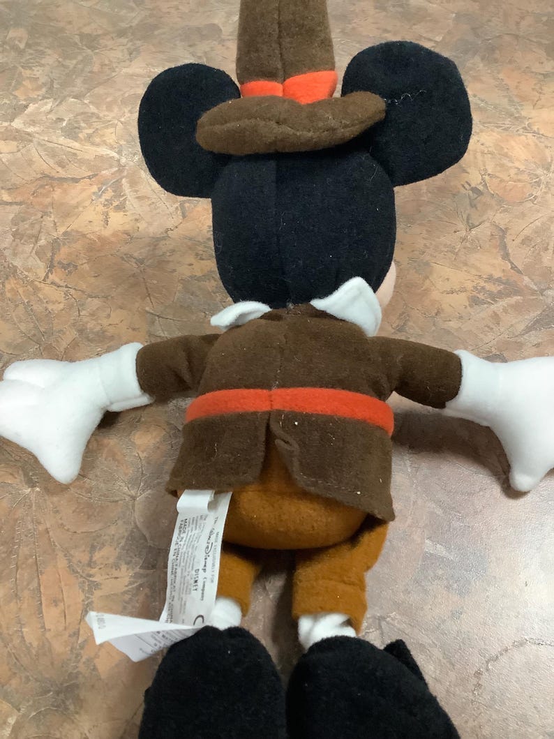 Puede incluir: Un peluche de Mickey Mouse con un sombrero y una chaqueta marr&oacute;n de peregrino con un cintur&oacute;n naranja. El juguete tiene orejas negras, una cabeza negra, guantes blancos y zapatos negros. El juguete est&aacute; sobre una superficie marr&oacute;n claro.