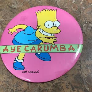 May include: A pink circular button featuring Bart Simpson from The Simpsons cartoon. Bart is depicted in his signature outfit, running with a mischievous grin. The text "AYE CARUMBA!" is displayed across the button.