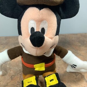 Puede incluir: Un peluche de Mickey Mouse vestido con un traje de peregrino marr&oacute;n con un sombrero a juego. La figura tiene orejas negras, cara rosa y guantes blancos. El atuendo incluye una hebilla de cintur&oacute;n y hebillas de zapatos amarillas.