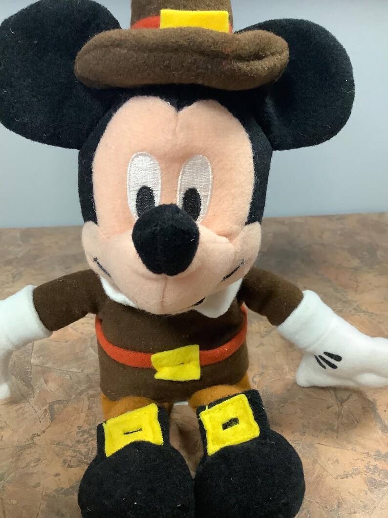 Puede incluir: Un peluche de Mickey Mouse vestido con un traje de peregrino marr&oacute;n con un sombrero a juego. La figura tiene orejas negras, cara rosa y guantes blancos. El atuendo incluye una hebilla de cintur&oacute;n y hebillas de zapatos amarillas.