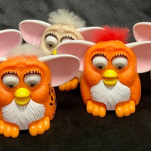 May include: Five colorful Furby toys. The toys are all in different colors, including purple, orange, yellow, and white. They have large, round eyes and fuzzy fur.