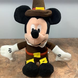 Puede incluir: Mu&ntilde;eco de peluche de Mickey Mouse vestido con un atuendo de peregrino marr&oacute;n, con sombrero, cintur&oacute;n y hebilla. El mu&ntilde;eco tiene orejas negras, cara blanca y guantes blancos. El mu&ntilde;eco mide unos 25 cm de alto.