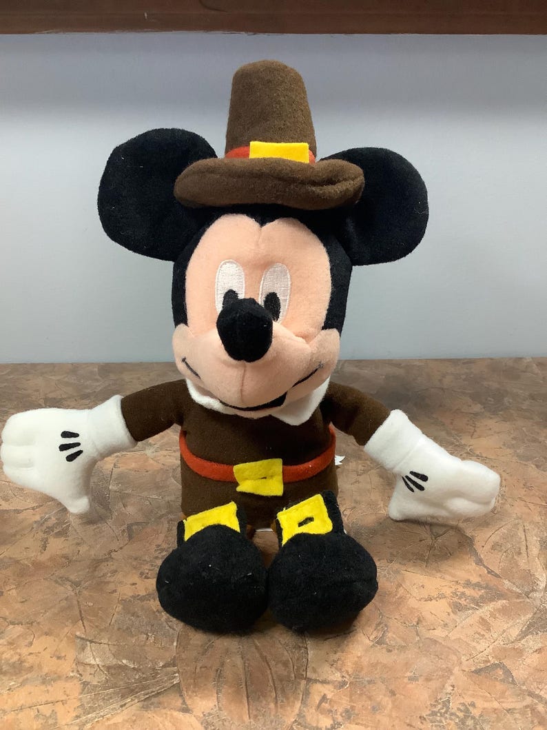 Puede incluir: Mu&ntilde;eco de peluche de Mickey Mouse vestido con un atuendo de peregrino marr&oacute;n, con sombrero, cintur&oacute;n y hebilla. El mu&ntilde;eco tiene orejas negras, cara blanca y guantes blancos. El mu&ntilde;eco mide unos 25 cm de alto.