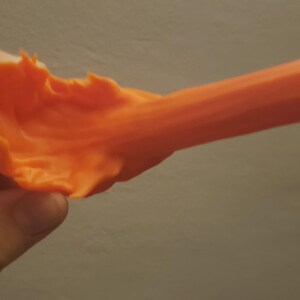 May include: Bright orange, pliable material held in a hand. The material has a textured, ruffled edge on one side and tapers into a smooth, elongated shape. The background is a neutral gray.