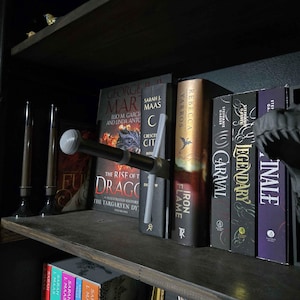 Könnte beinhalten: Ein Bücherregal mit verschiedenen Büchern und dekorativen Gegenständen. Die Bücher haben farbenfrohe Buchrücken und Titel wie "Iron Flame" und "Caraval". Ein dekoratives Schwert liegt auf den Büchern, und kleine Vogelfiguren befinden sich auf dem obersten Regal.