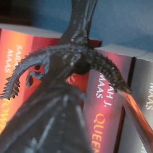 May include: A black dragon figurine with a spiky tail and wings, perched on top of a bookshelf with several books. The books are all by Sarah J. Maas and have the titles "Throne of Glass", "Crown of Midnight", "Heir of Fire", "Queen of Shadows", and "A Court of Thorns and Roses".