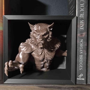 Könnte beinhalten: Braune Werwolf-Skulptur, die aus einer schwarzen Rahmung hervortritt. Die detaillierte Figur hat einen muskulösen Torso, spitze Ohren und einen grimmigen Ausdruck. Das Kunstwerk wird auf einem Holzregal neben einem Buch ausgestellt.