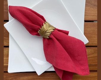 Könnte beinhalten: Eine rote Leinen-Serviette wird von einem goldfarbenen, blattförmigen Serviettenring gehalten und auf zwei weißen quadratischen Tellern platziert. Die Teller stehen auf einem Holztisch, was einen Kontrast von Farben und Texturen erzeugt.