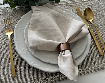 Könnte beinhalten: Eine beigefarbene Leinen-Serviette, die zu einem Dreieck gefaltet und auf einen weißen Teller gelegt wird. Ein brauner Serviettenring hält die Serviette zusammen. Der Teller liegt auf einem silbernen Platzdeckchen.