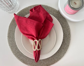Könnte beinhalten: Eine rote Leinen-Serviette, in Rosenform gefaltet, liegt auf einem weißen Teller. Ein gewebter, cremefarbener Serviettenring hält die Serviette. Der Teller steht auf einem Perlen-Platzset. Ein Glas und eine Kerze sind ebenfalls im Bild.