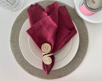 Könnte beinhalten: Eine burgunderfarbene Leinen-Serviette, gefaltet und von einem Serviettenring aus Naturwebstoff gehalten. Die Serviette liegt auf einem weißen Teller, auf einem Perlen-Charger. Eine Kerze und ein Glas sind im Hintergrund.