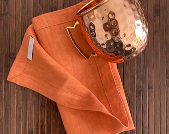 Könnte beinhalten: Eine orangefarbene Leinen-Serviette mit einem gehämmerten Kupferbecher darauf. Der Becher hat einen goldenen Griff und steht auf einer Bambusoberfläche.