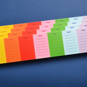 May include: A set of six colorful weekly planner pads with days of the week labeled in black text. The pads are stacked on top of each other, with the yellow pad on the bottom and the pink pad on top. The pads are lined with white lines for writing.