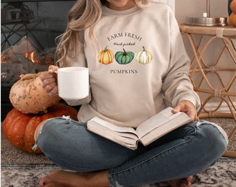 May include: A woman wearing a beige sweatshirt with the text "Farm Fresh Hand Picked Pumpkins" and a graphic of three pumpkins. She is sitting on a rug with a book in her lap and a mug in her hand.