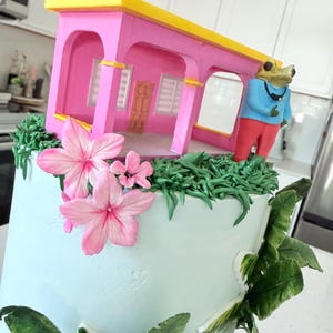 Puede incluir: Un pastel azul claro decorado con hierba de glaseado verde, flores rosas y hojas de color verde oscuro. Encima hay una estructura de casa rosa con techo amarillo, acompañada de una pequeña figura de rana con camisa azul y pantalones rojos.