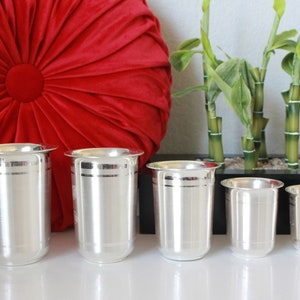 May include: Five silver drinking glasses of varying sizes, with a decorative band around the middle of each glass. The glasses are arranged in a row on a white surface.