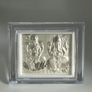 May include: A silver framed picture of two Hindu deities, Lakshmi and Ganesha, in a clear plastic case. The picture is embossed with the deities' names in Hindi.
