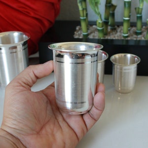 May include: Three silver drinking glasses with a decorative band around the middle. The glasses are made of polished metal and have a shiny finish.