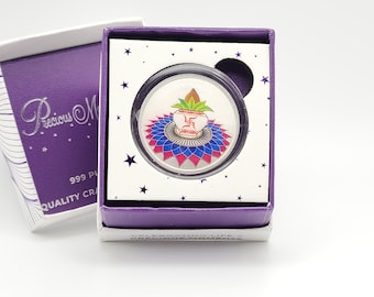 May include: A silver coin with a colorful design of a pot with a flame and a swastika symbol. The coin is in a purple box with stars and the text "Precious Moments" and "Celebrating Life's Moments".