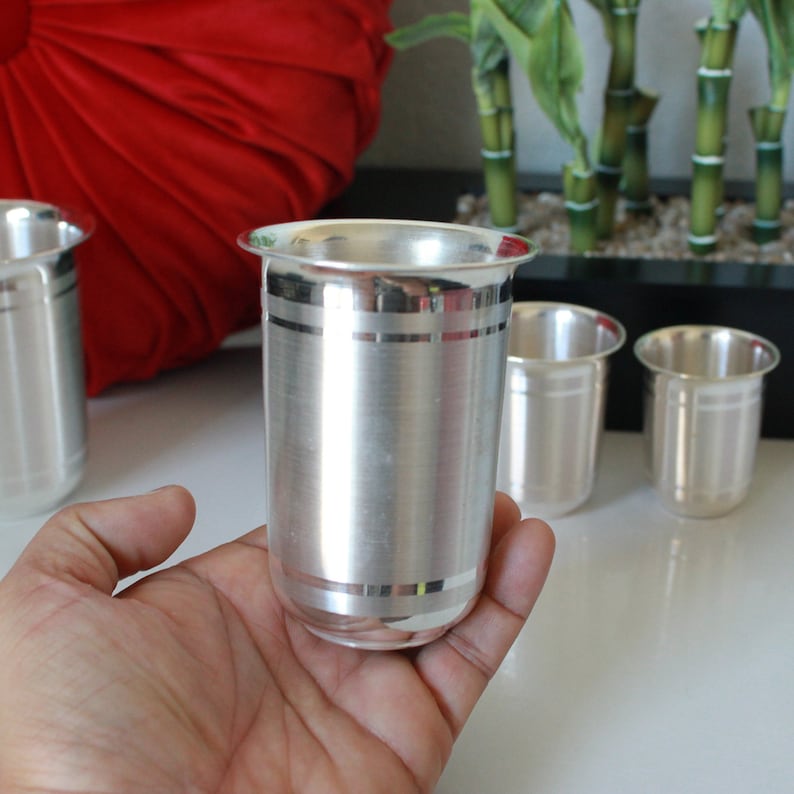 May include: Four silver drinking glasses with a decorative band around the middle. The glasses are on a white surface.